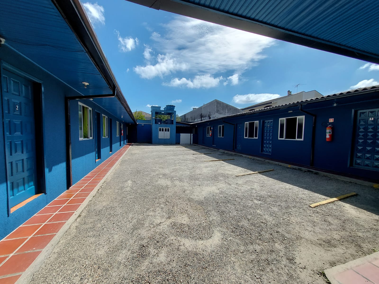 Pátio privado do motel com estacionamento e portas azuis das suítes